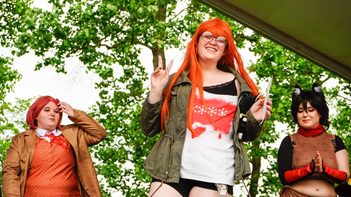 Three cosplayers stand on an outdoor stage, one center in an orange wig and glasses flashing a peace sign, flanked by a red-wig schoolgirl cosplayer and a cat-eared cosplayer at Knox Pop Con.